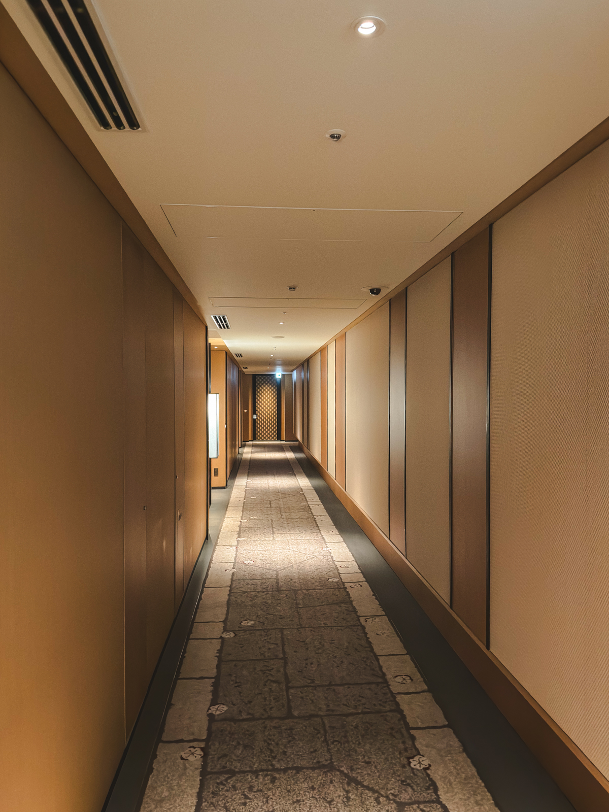 Long hotel hallway with beige walls and some light fixtures on the side.