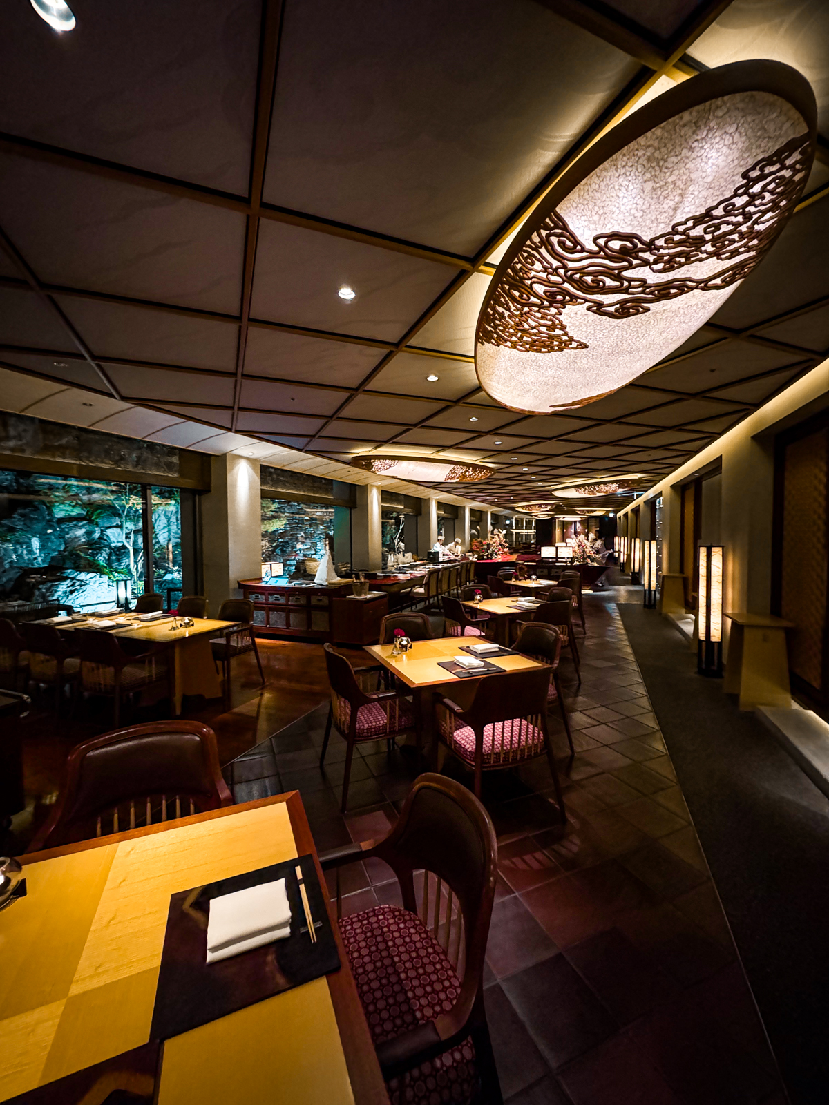 Mizuki main dining area with square tables and a long sushi bar in the distance with soft lighting and dark tiles.