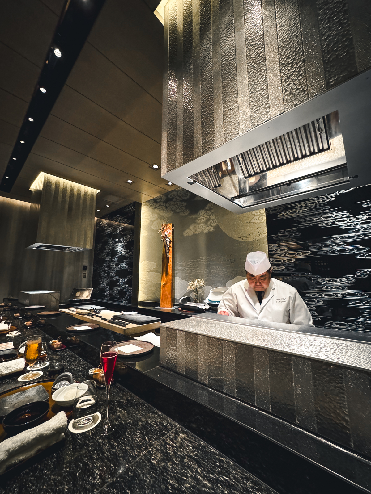 Bar seating counter with chef Takashi-sama behind the counter with two large smoke hoods and black and silver decor. 