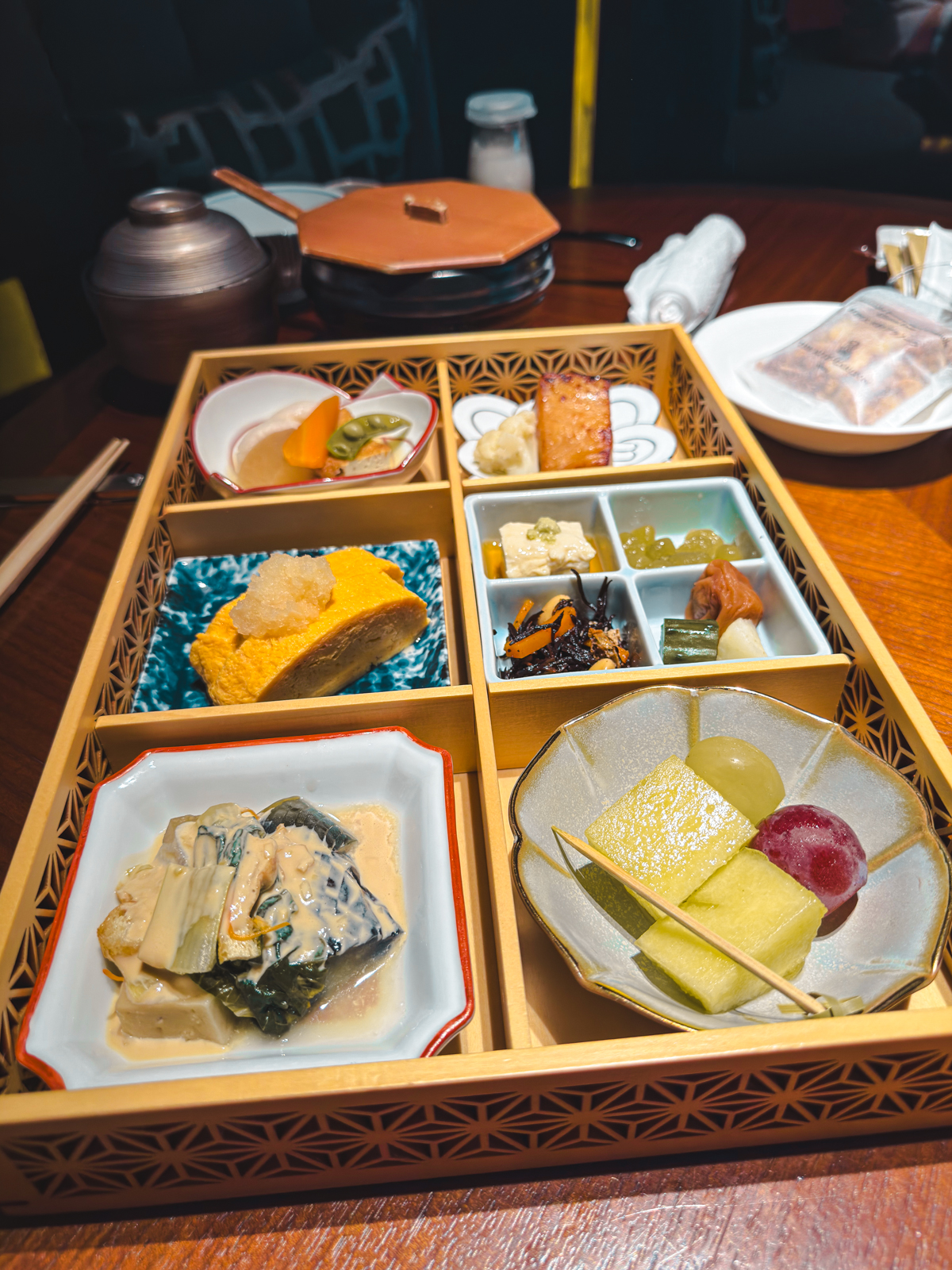 Japanese set menu box with 6 squares featuring fruits, egg, pickles, grilled fish, tofu, and congee bowl.