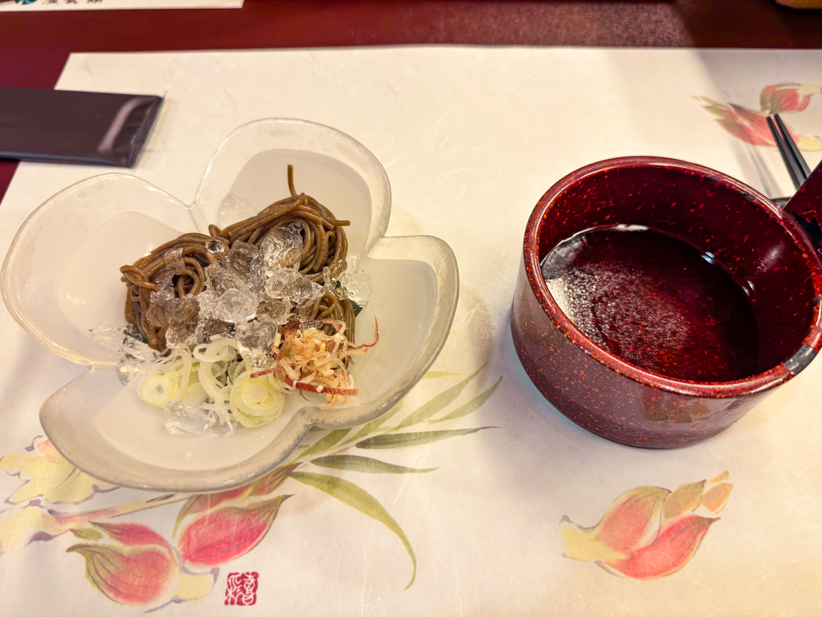 Cold soba noodles with diced onions, green onions, and ice cubes with a small bowl of dashi on the side.